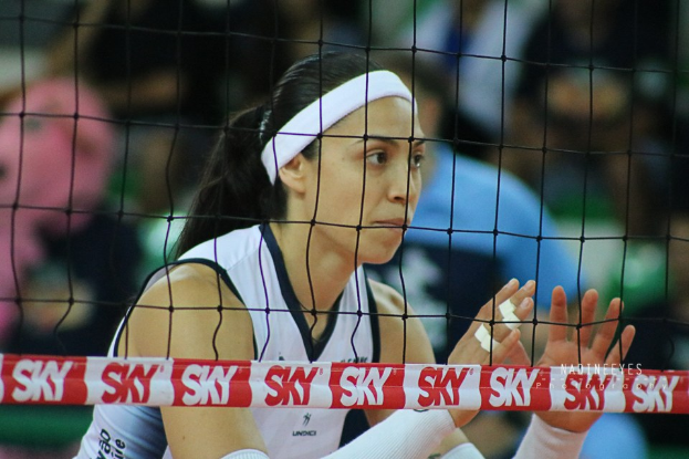 Eine weibliche Volleyballspielerin in Uniform springt in der Nähe des Netzes mit konzentrierter Miene, ihr unscharfer Hintergrund deutet auf Spielaktion hin.