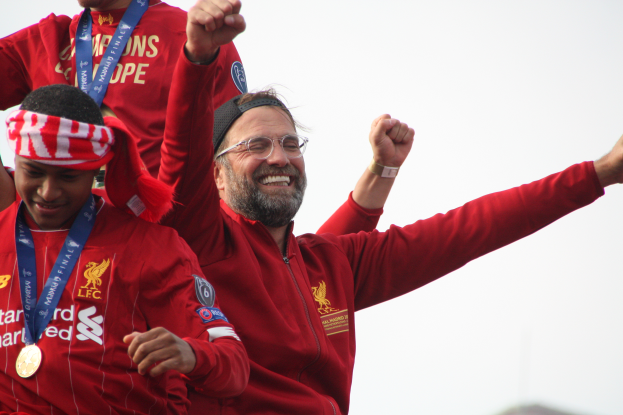 Liverpool-Trainer Jürgen Klopp feiert mit seinen Spielern nach dem Gewinn der UEFA Champions League, während zwei medalletragende Personen in der Nähe sind und der Himmel im Hintergrund zu sehen ist.