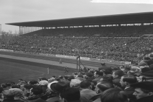 Schwarzes und weißes Foto eines vollen Stadions mit Zuschauern, die ein Fußballspiel verfolgen, mit Bannern, Pfählen, einem Schuppen, Bäumen, einem Turm und einer bewölkten Himmel