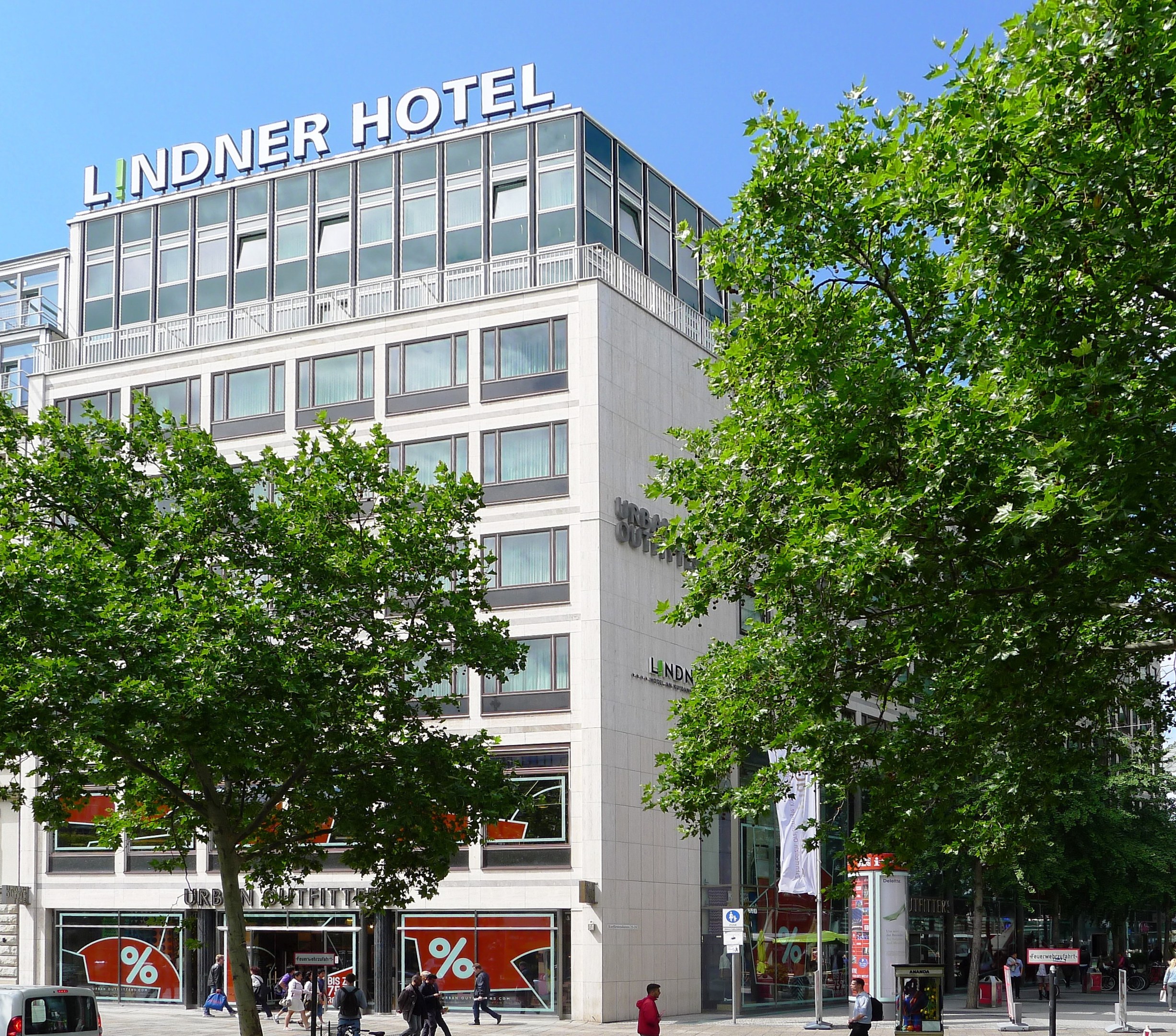 Großes modernes Hotelgebäude mit Glasfenstern und Beschriftung oben, umgeben von Bäumen und einer belebten Straße mit Fußgängern und Fahrzeugen, unter einem sichtbaren Himmel.