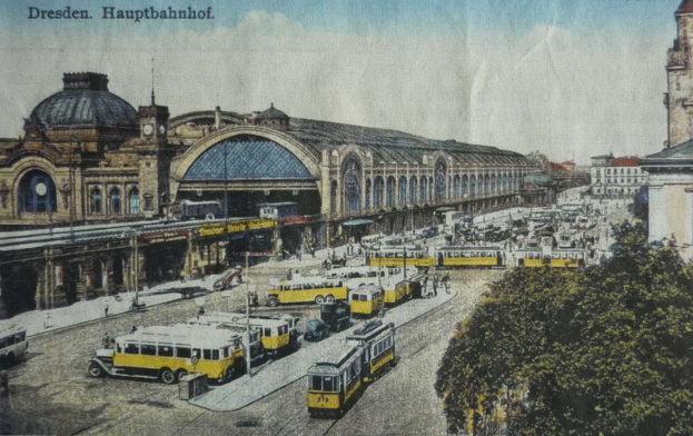Eine alte Postkarte, die einen belebten Dresdner Bahnhof mit Fahrzeugen, Menschen, Bäumen, Gebäuden und einem bewölkten Himmel zeigt, mit der Aufschrift "Dresden Hauptbahnhof" oben.