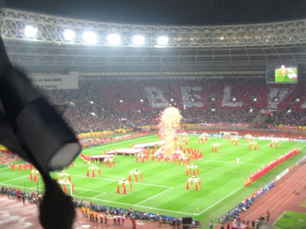 Ein Stadion mit Flutlicht, Eisengitter, Zuschauern, stehenden Personen, Werbetafeln, Anzeigetafeln, Überwachungskamera, Flaggen und Dekorationselementen.