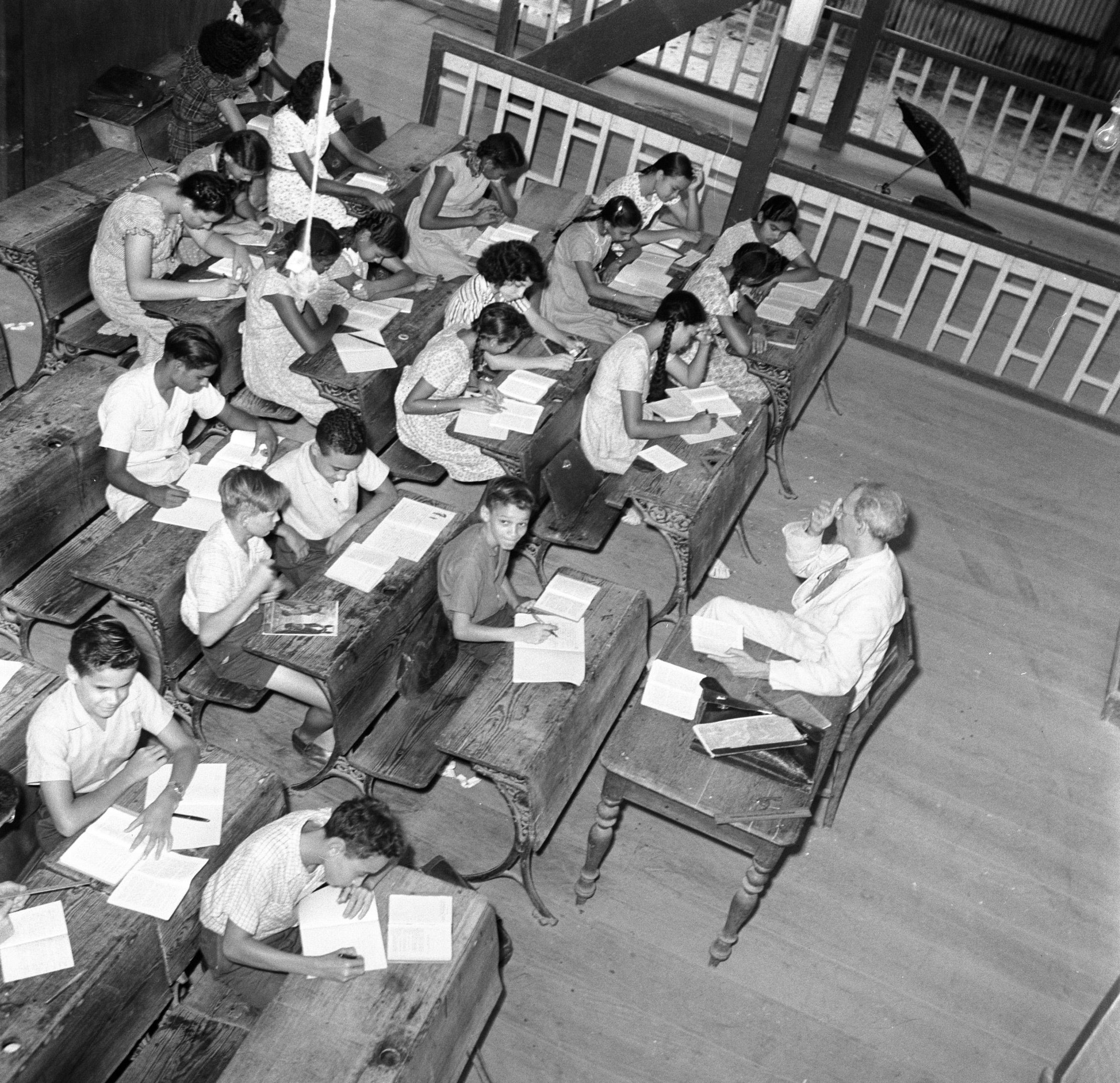 Schwarzes und weißes Foto eines Klassenzimmers mit Kindern an Tischen, Büchern und Gegenständen auf den Tischen und einem Geländer und einem Schirm im Hintergrund.