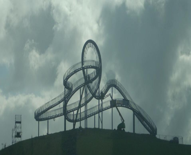 Eine Achterbahn auf dem Höhepunkt eines Hügels unter einem bewölkten Himmel, umgeben von Stützkonstruktionen.