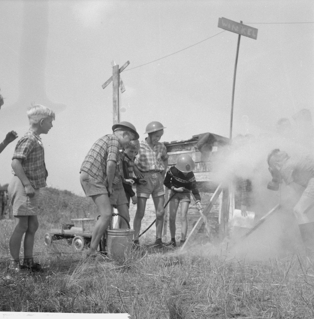 Schwarzes Bild von Kindern in Helmen, die sich um einen Hydranten in einem grasbewachsenen Feld versammeln, einige halten Gegenstände, mit Strommasten und einem Schild im Hintergrund.