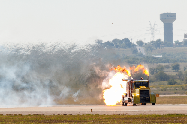 Ein gelber Lkw fährt auf einer Straße mit großen Flammen, umgeben von Gras, mit Bäumen, Türmen, Dröhnen und einem klaren blauen Himmel im Hintergrund.