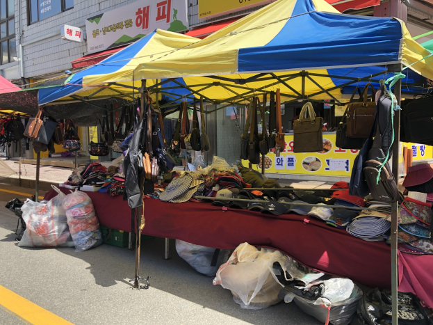 Ein Straßenmarkt mit Zelten, auf denen Taschen, Hüte und andere Gegenstände präsentiert werden, vor Häusern mit Fenstern und Texttafeln.
