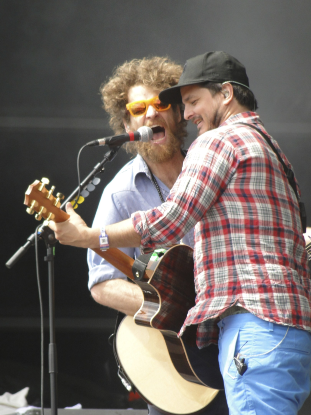 Zwei Männer mit Gitarren und Mikrofonen auf einer Bühne, einer trägt ein rot-blau kariertes Hemd mit blauen Hosen, der andere eine Sonnenbrille und eine schwarze Mütze.