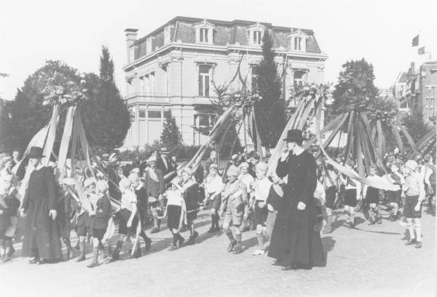 Schwarzes und weißes Bild einer Gruppe von Menschen, die eine Straße entlanggehen und Fahnen halten, mit Hüten, Bäumen und Gebäuden im Hintergrund, das einen Maypole zeigt.