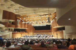 Eine große Halle voller Menschen, die ein Konzert verfolgen, mit einer Gruppe auf der Bühne, die Instrumente spielt, und anderen, die auf Stühlen sitzen, unter einer von Lichtern geschmückten Decke.