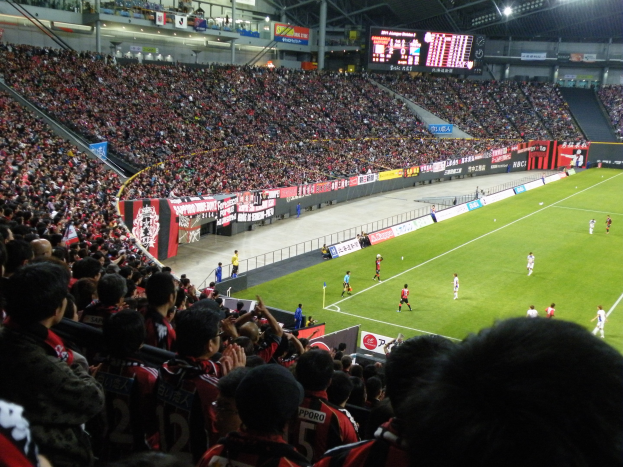 Ein Fußballspiel in einem großen Stadion mit Spielern auf dem Feld und Zuschauern auf den Rängen, mit Bannern, einem Zaun, einem Display, Metallstangen und Deckenleuchten.