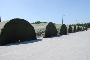 Eine Reihe von Militärzelten auf einem Parkplatz mit einem Feuerlöscher davor, Straßenlaternen, Bäumen und einem klaren blauen Himmel im Hintergrund.
