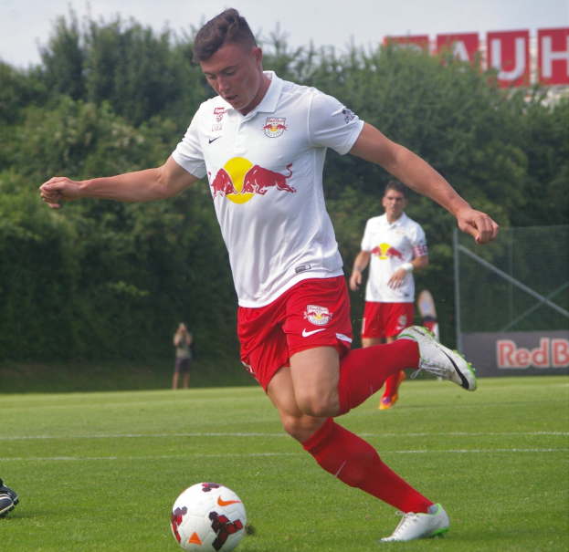 Ein professioneller Fußballspieler in einem roten Trikot kickt einen Ball auf einem grünen Feld mit Bäumen und einem klaren blauen Himmel im Hintergrund, wobei ein "RB Leipzig"-Schild sichtbar ist.