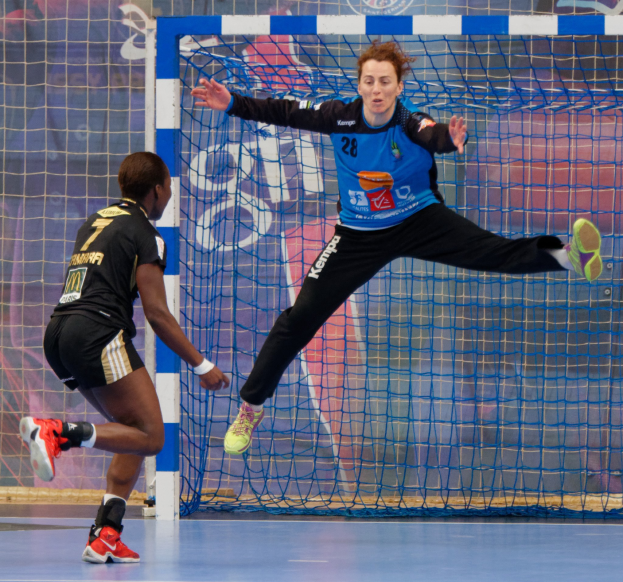 Zwei Frauen spielen Handball auf einem Platz mit einem Tor und einer Fahne im Hintergrund, beide tragen sportliche Schuhe.