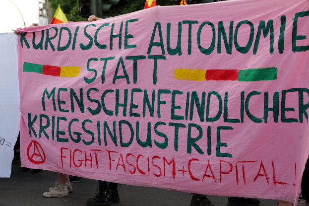 Eine Gruppe von Menschen mit einer pinken Fahne mit der Aufschrift 'Kurdische Autonomie Statt Menschenfeindlicher Kriegsindustrie Fight Fascism Capital' auf einer Straße mit Ampeln, Flaggen, Bäumen, Gebäuden und einem klaren blauen Himmel im Hintergrund.