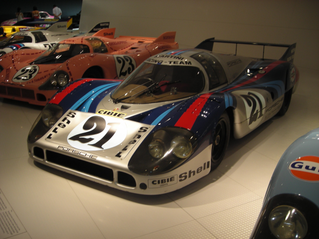 Porsche 917K Rennwagen in einem Museum ausgestellt, umgeben von anderen Sportwagen, mit Menschen drumherum und einer Wand im Hintergrund; ein Blatt Papier mit Text ist links sichtbar.
