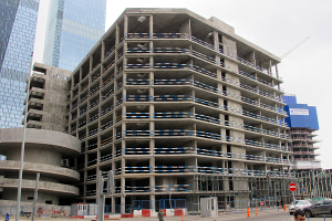 Ein großes Bauwerk im Bau in einer Stadt, umgeben von Wolkenkratzern, Fahrzeugen, Fußgängern und verschiedenen städtischen Elementen unter einem klaren blauen Himmel.