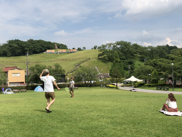 Gruppe von Menschen, die Badminton in einem Park spielt, mit einem Mann, der einen Schläger hält und auf Gras in der Nähe von Zelten und Gebäuden sitzt, mit Bäumen, Hügeln und einem bewölkten Himmel im Hintergrund.