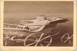 Ein altes Schwarz-Weiß-Foto eines Babys in einem hölzernen Kinderwagen mit vier Rädern, das eine weiße Strampelanzug trägt und einen friedlichen Ausdruck hat, mit kursiver Schrift am unteren Rand.