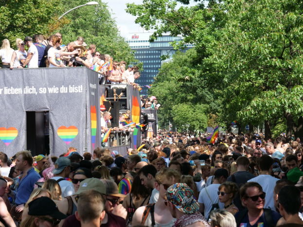Eine große Menschenmenge steht vor einem Lastwagen bei der Christopher Street Day Parade in Berlin, viele tragen Mützen und Schutzbrillen, einige halten Fahnen, mit einem Banner auf dem Lastwagen und Bäumen, Gebäuden und einem Laternenpfahl im Hintergrund bei bewölktem Himmel.