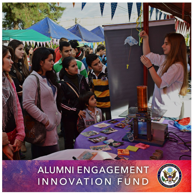 Gruppe von Menschen um einen Tisch versammelt mit einer "Alumni Engagement Innovation Fund"-Aufschrift, umgeben von Gegenständen, mit Zelten und Bäumen im Hintergrund unter einem klaren blauen Himmel und Text mit einem Logo unten.