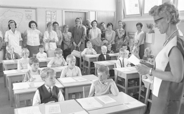 Schwarz-weißes Foto eines Lehrers, der vor einem Klassenzimmer mit Kindern auf Bänken und auf dem Boden sitzend und stehend steht und Papiere hält, während Papiere und Bücher auf Tischen liegen, mit einer Tafel, Fenstern und einer Blumenvase im Hintergrund.