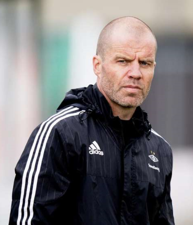 Ein kahlköpfiger Mann in einer schwarzen Jacke steht auf einem Fußballfeld und trägt eine marineblaue Jacke mit weißen Streifen und einem weißen Kragen, die Hände sind verschränkt und er hat einen ernsten Gesichtsausdruck.