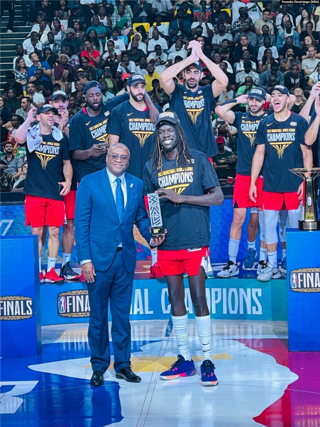 Eine Gruppe von Männern auf einem Basketballfeld hält Trophäen, während Menschen im Hintergrund sitzen; zwei Trophäen ruhen auf Podesten rechts, was auf einen Sieg in den NBA-Finals hinweist.