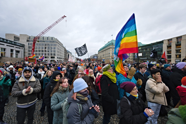 Eine große Gruppe von Menschen mit Fahnen und Schildern vor einem Gebäude während einer LGBTQ+-Rechtsdemo in Berlin, mit Gebäuden, einem Kran und einem bewölkten Himmel im Hintergrund.