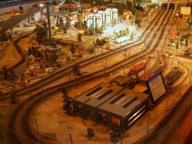 Eine detaillierte Modellbahnausstellung in einem Museum, die Gebäude, Bäume, Eisenbahnschienen, Fahrzeuge, Pfähle, Lichter, Platten und andere Gegenstände umfasst, die eine belebte Stadtlandschaft nachahmen.