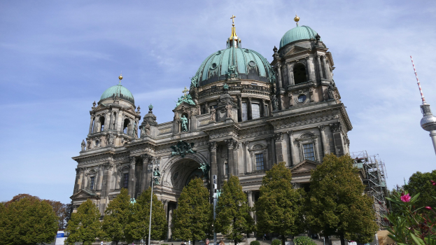 Berliner Dom, eine große Kathedrale mit Fenstern, Säulen, Bögen und Statuen, vor einem bewölkten Himmel, mit Menschen, Bäumen und einem Turm im Vordergrund.