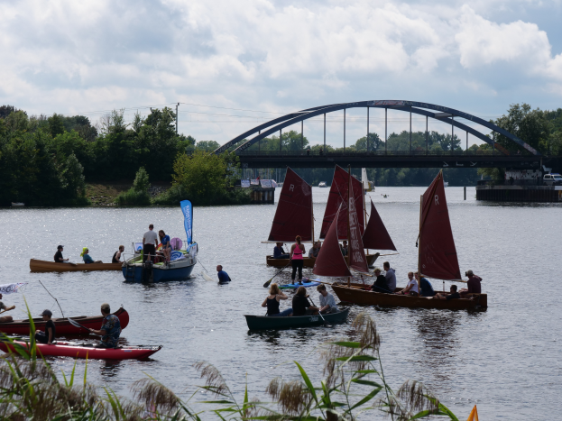 Eine Gruppe von Menschen in kleinen Booten auf einem Gewässer, im Hintergrund eine Brücke, Fahrzeuge, Bäume und ein bewölkter Himmel, sowie Pflanzen unten, die auf eine Regatta hindeuten.