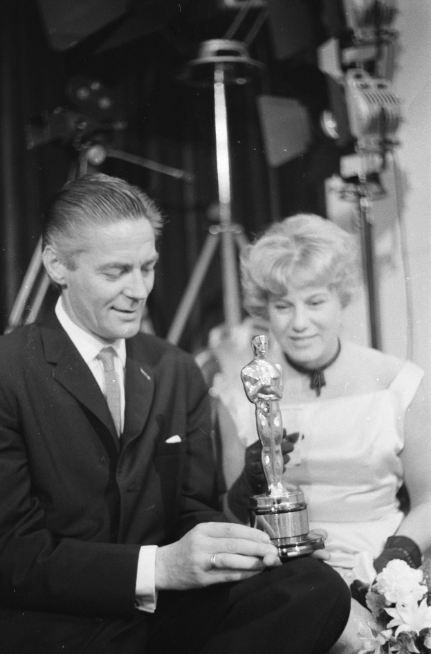 Ein Mann in einem schwarzen Blazer, einem weißen Hemd und einer Krawatte h├Ąlt eine Oscar-Statue in der Hand, waehrend er neben einer Frau in Abendgarderobe sitzt, die einen Blumenstrauss h├Ąlt, mit weicher Beleuchtung im Hintergrund.