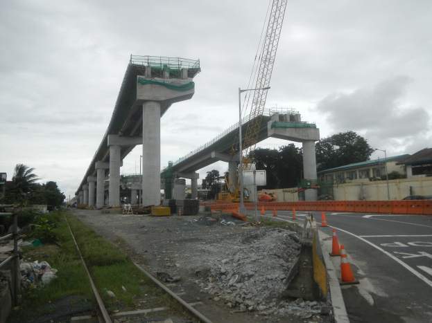 Baustelle mit einer Brücke im Hintergrund, Straße durch Verkehrskegel markiert, Bahnschiene links, verstreute Steine und Gras, Bäume und Gebäude säumen die Straße und ein bewölkter Himmel.