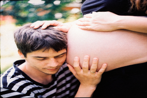 Ein Mann und eine Frau stehen nebeneinander, der Mann berüttend die Frau's Bauch und schaut auf ihr ungeborenes Kind.