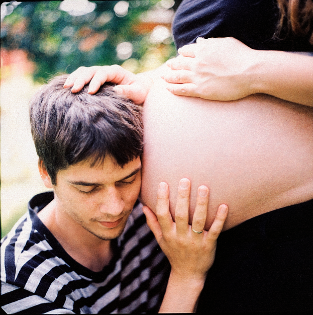 Ein Mann und eine Frau stehen nebeneinander, der Mann berüttend die Frau's Bauch und schaut auf ihr ungeborenes Kind.