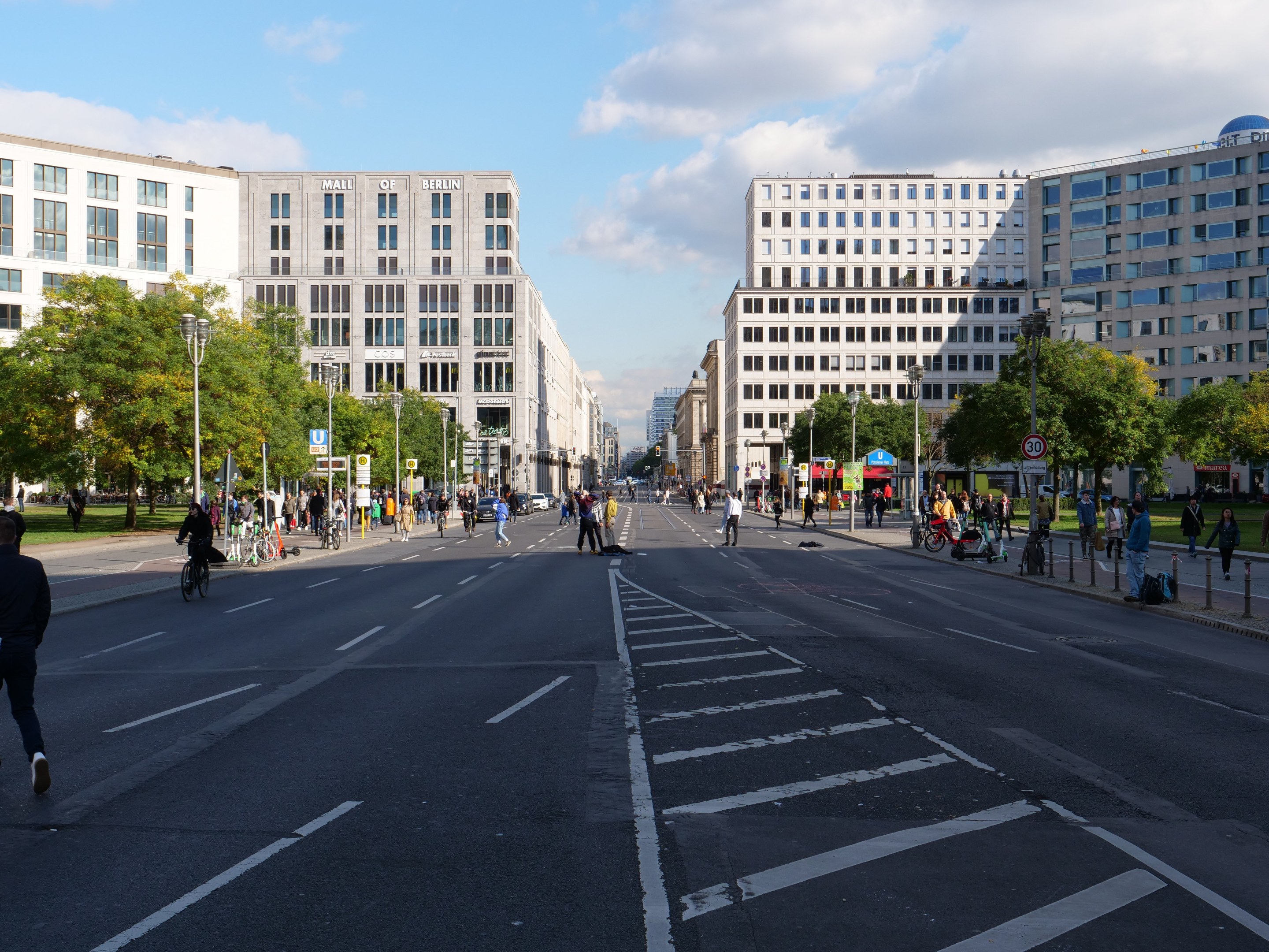Eine belebte Stadtstraße in Berlin, Deutschland, mit Fußgängern und Radfahrern auf der Straße, umgeben von hohen Gebäuden, Bäumen, Laternenmasten und Schildern unter einem bewölkten Himmel.