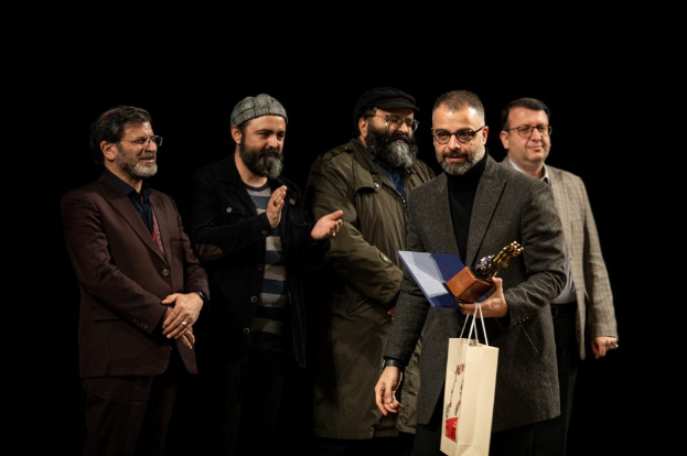 Eine Gruppe von Männern steht auf einer Bühne zusammen, einer hält eine Tasche und eine Trophäe, mit einem dunklen Hintergrund, der die Eröffnung der Berliner Filmfestspiele suggeriert.