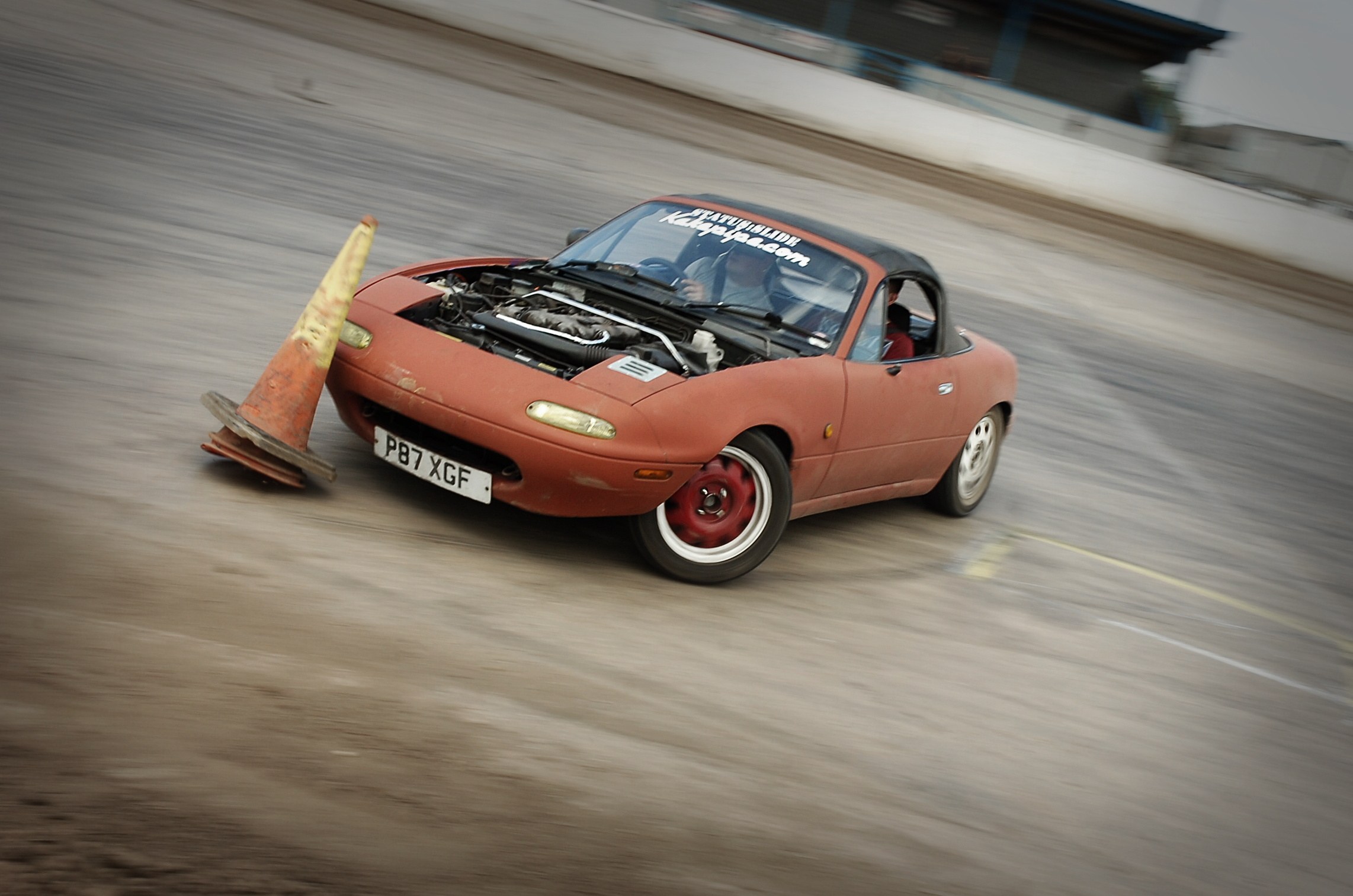 Eine Person, die ein rotes Auto auf einer Rennstrecke fährt, mit einem Verkehrskegel vorneweg, der unscharfe Hintergrund deutet auf hohe Geschwindigkeit hin.