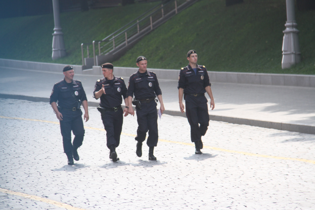 Drei Polizisten in Uniform und Mützen gehen eine Straße entlang mit Hintergrundelementen wie Pfosten, Geländern, Treppen und Gras.
