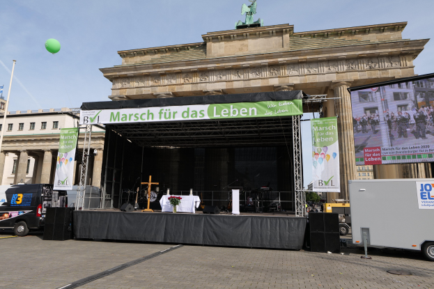 Bühne vor dem Brandenburger Tor in Berlin aufgebaut, mit einem Tisch mit einer Blumenvase, einem Kreuzsymbol, Bannern, Lautsprechern und anderen Gegenständen, sowie Fahrzeugen auf der Straße und Gebäuden im Hintergrund.