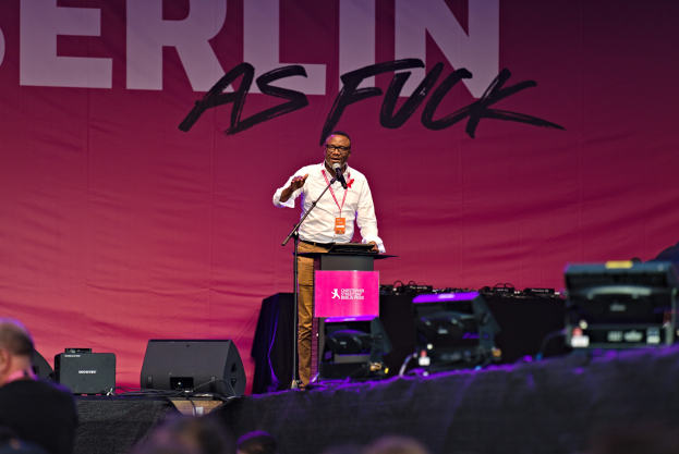 Ein Mann in weißem Hemd und braunen Hosen steht auf einer Bühne und hält ein Mikrofon, mit einem pinken Banner 'Berlin as Fuck' im Hintergrund und einer Menge davor.