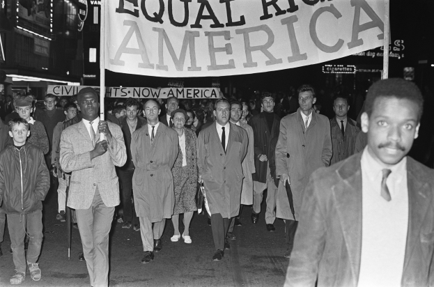 Schwarzes und weißes Foto einer Gruppe von Menschen, die eine Straße entlangmarschieren und ein Banner mit der Aufschrift "Gleiche Rechte Amerika" tragen, mit Gebäuden und Straßenlaternen am Straßenrand.
