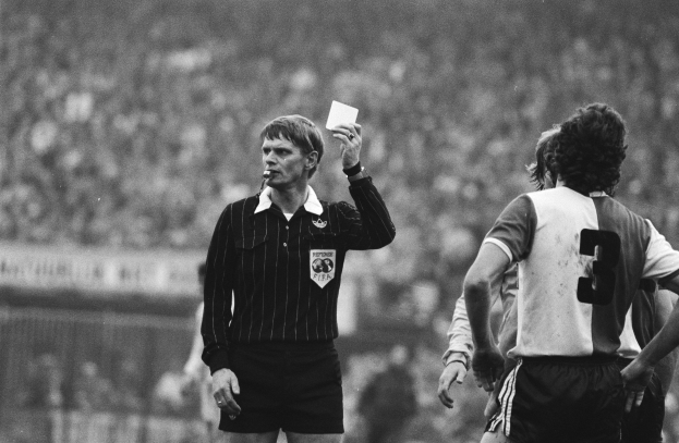 Schwarzes und weißes Bild eines Schiedsrichters mit roter Karte, mit einer Menge von Menschen in Fußballuniformen hinter einem Zaun.