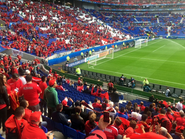Eine große Menschenmenge in einem hell erleuchteten Stadion schaut einem Füßtballspiel zu, mit Zuschauern auf Sitzen und Stehplätzen, sichtbaren Bannern und einem Tor im Hintergrund.