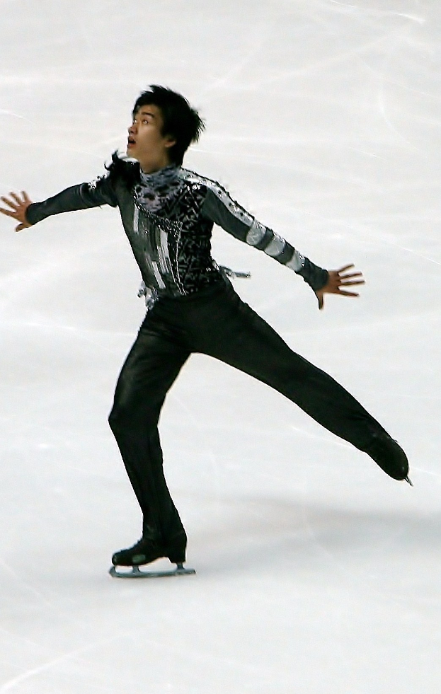 Männlicher Eiskunstläufer in einem schwarzen Kleid, der eine Choreografie auf einem Eisstadion präsentiert