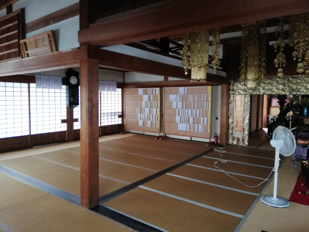 Traditionelles japanisches Zimmerinterieur mit Tatami-Matten, Tischventilator, Teppich, Feuerlöscher, Gegenständen auf Oberflächen, Uhr, Wandpapiere, hölzernen Pfählen, Deckenleuchten und einem Kronleuchter.