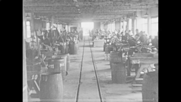Schwarzes und weißes Foto einer Fabrik mit Arbeitern, die Maschinen bedienen, nummerierten Fässern, einer Eisenbahnschiene, Säulen und Deckenlampen während der industriellen Revolution.