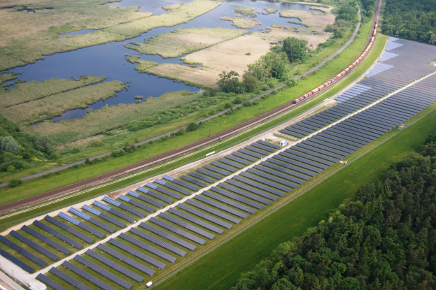 Luftaufnahme einer Solar-Farm mit Solar-Panelen auf einem Feld, umgeben von Bäumen, Gras, Pflanzen und Wasser, mit einem Zug auf einer nahen Eisenbahnstrecke.