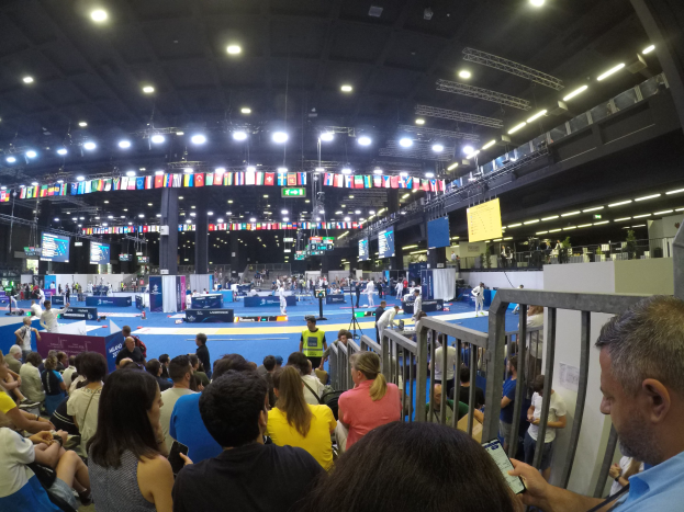 Eine große Menschenmenge auf Sitzplätzen und Stehplätzen in einer Indoor-Sportarena mit Bildschirmen, Fahnen und Deckenbeleuchtung im Hintergrund.